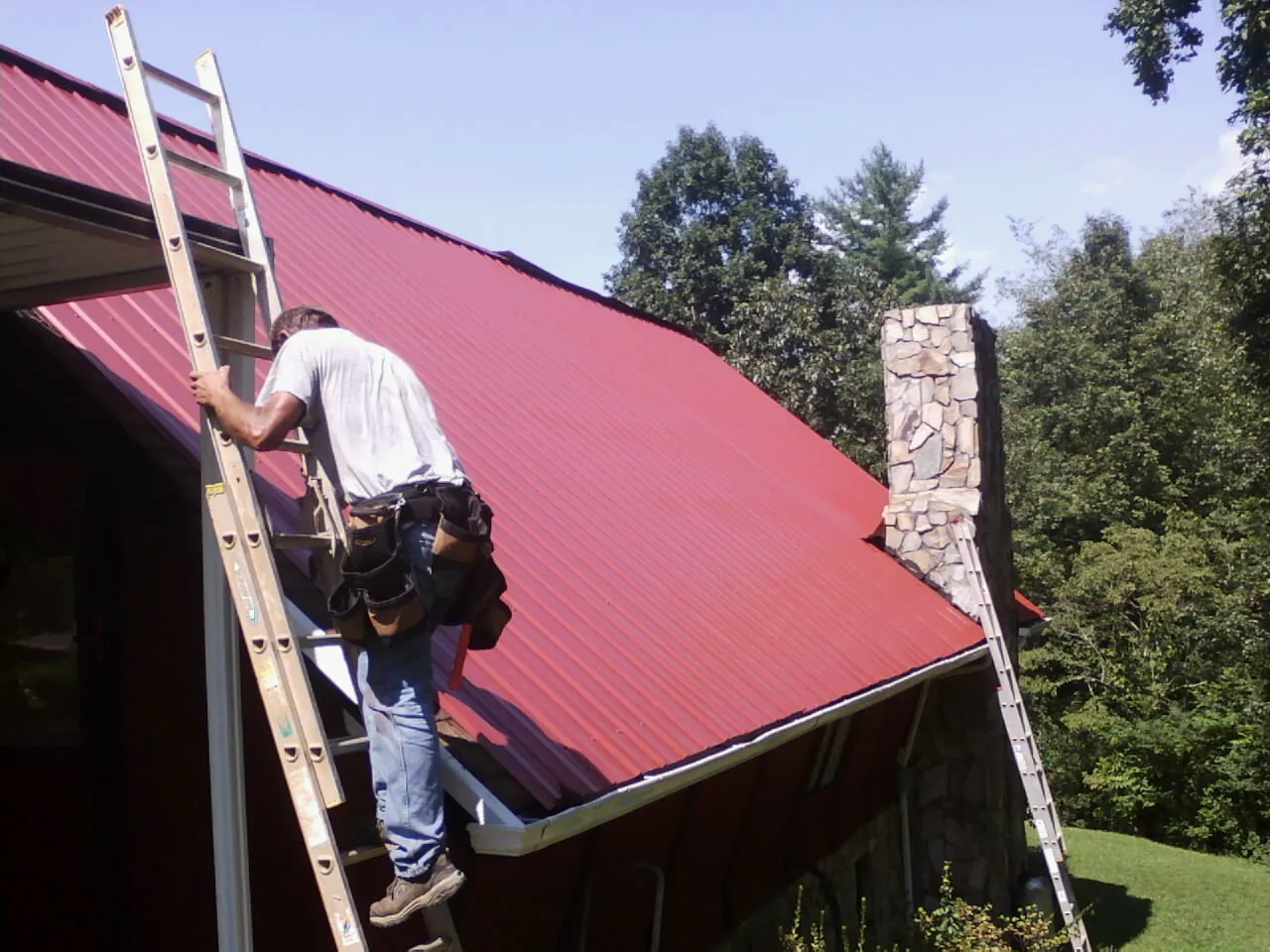 Roofer on a ladder beside a red metal roof and stone chimney — Soffit & Fascia Repair by Roofing Brothers in San Miguel